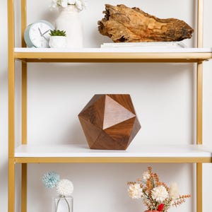 May include: A gold metal shelf unit with white shelves. The middle shelf has a dark brown geometric wooden sculpture. The top shelf has a white vase with white flowers, a small green plant in a white pot, and a white clock.