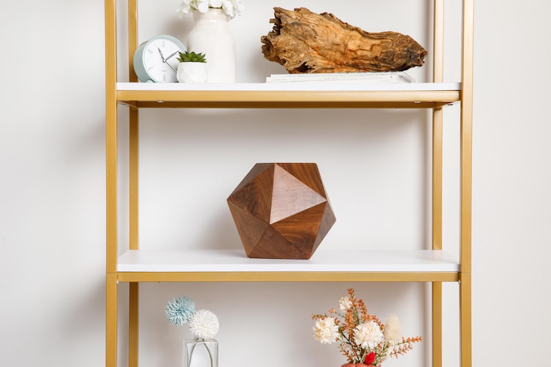 May include: A gold metal shelf unit with white shelves. The middle shelf has a dark brown geometric wooden sculpture. The top shelf has a white vase with white flowers, a small green plant in a white pot, and a white clock.