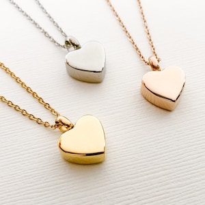 May include: Three heart-shaped cremation necklaces, one silver, one gold, and one rose gold. Each necklace has a delicate chain and a heart-shaped pendant.