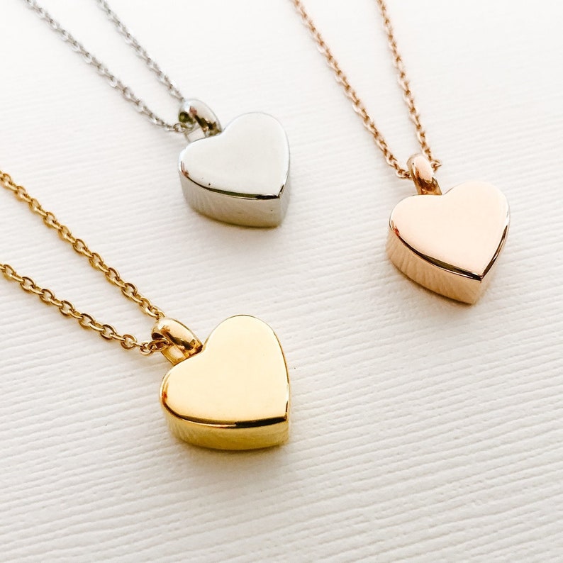 May include: Three heart-shaped cremation necklaces, one silver, one gold, and one rose gold. Each necklace has a delicate chain and a heart-shaped pendant.