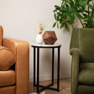 May include: A round white marble end table with a black metal frame. The table has a wooden geometric sculpture and a white ceramic vase with dried flowers on top. A brown leather sofa and a green velvet armchair are in the background.