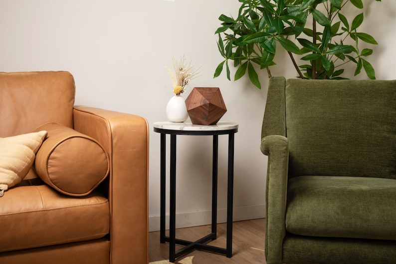 May include: A round white marble end table with a black metal frame. The table has a wooden geometric sculpture and a white ceramic vase with dried flowers on top. A brown leather sofa and a green velvet armchair are in the background.