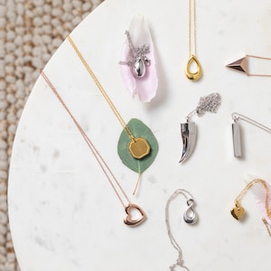May include: Assortment of silver, gold, and rose gold necklaces with various pendant shapes, including a heart, infinity symbol, teardrop, and geometric shapes. The necklaces are displayed on a white marble surface with pink and green accents.