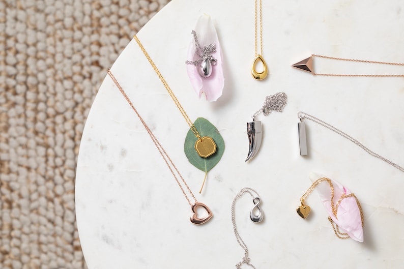 May include: Assortment of silver, gold, and rose gold necklaces with various pendant shapes, including a heart, infinity symbol, teardrop, and geometric shapes. The necklaces are displayed on a white marble surface with pink and green accents.