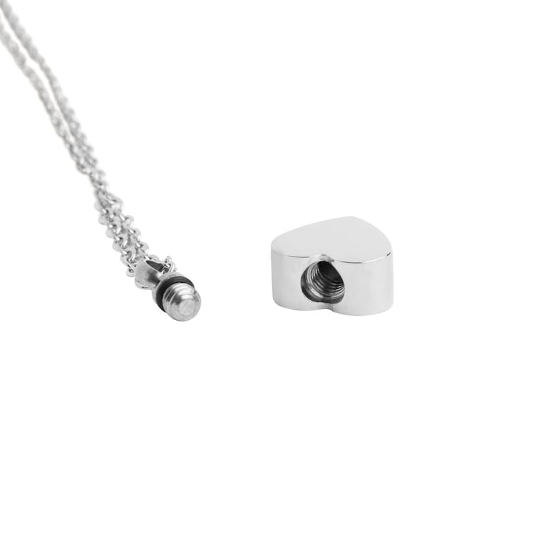 May include: A silver heart-shaped cremation necklace with a threaded screw closure. The necklace is on a silver chain.