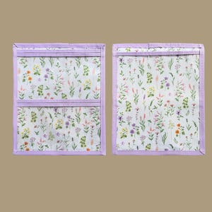 Pu&ograve; includere: Due organizer in tessuto rettangolari con sfondo bianco e una stampa colorata di fiori di campo. Gli organizer sono rifiniti con bordi in tessuto viola chiaro e includono strisce orizzontali in tessuto viola chiaro sui pannelli frontali.