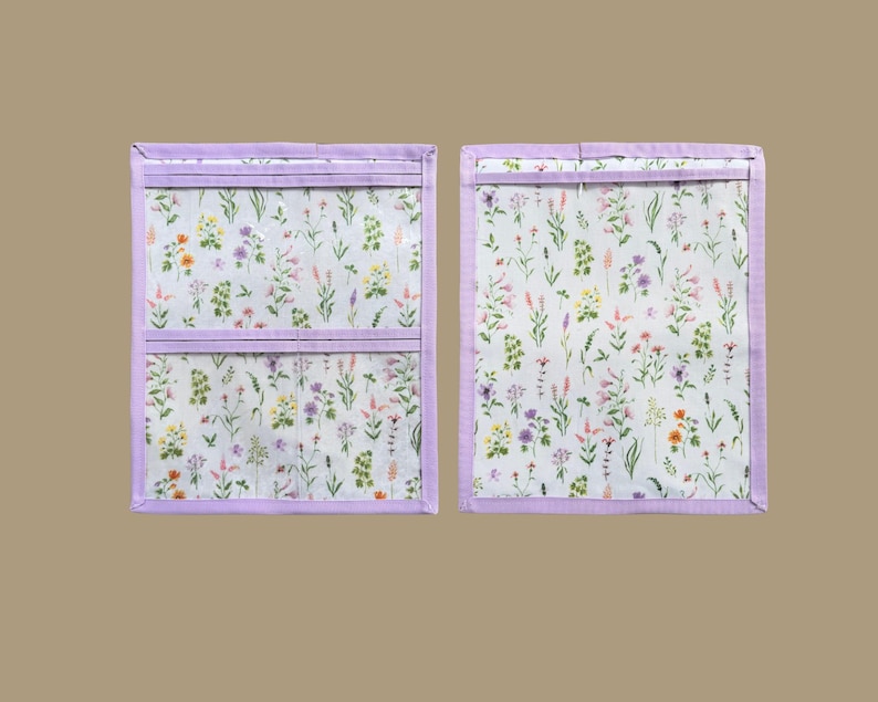 Pu&ograve; includere: Due organizer in tessuto rettangolari con sfondo bianco e una stampa colorata di fiori di campo. Gli organizer sono rifiniti con bordi in tessuto viola chiaro e includono strisce orizzontali in tessuto viola chiaro sui pannelli frontali.