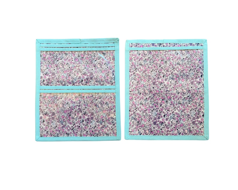 Pu&ograve; includere: Due organizer rettangolari in tessuto con bordo azzurro e un fitto motivo floreale con fiori rosa, viola e bianchi. Un organizer presenta due tasche orizzontali in plastica trasparente, l'altro ha un'unica grande tasca trasparente.