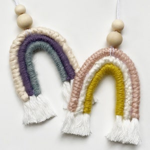 May include: Two colorful macrame rainbows with white tassels and wooden beads. The rainbows are made with different colored yarn, including purple, blue, pink, yellow, and white.