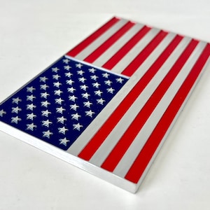 May include: A rectangular metal object featuring the American flag design. The flag has a blue field with white stars and red and white stripes. The object has a silver border and is set against a white background.