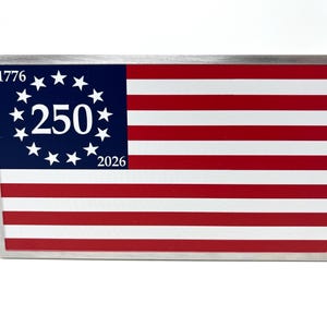 Puede incluir: Un letrero rectangular de metal con el diseño de la bandera estadounidense. La bandera tiene rayas rojas y blancas y un cantón azul con estrellas blancas y los números "250", "1776" y "2026".