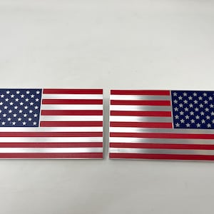 May include: Two rectangular metal plaques featuring the American flag design. Each plaque has a blue field with white stars and red and silver stripes. The plaques are displayed side by side.