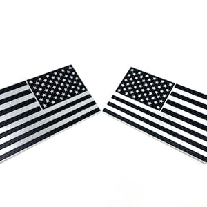 May include: Two black and silver American flag decals. Each decal features a field of white stars in the upper left corner and alternating black and silver stripes. The decals are rectangular and have a glossy finish.