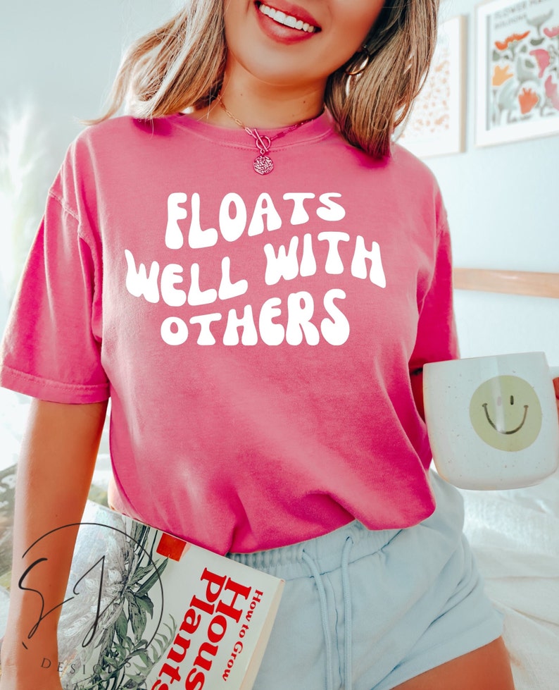 May include: A pink t-shirt with the text "FLOATS WELL WITH OTHERS" in white, held by a person. The person is also holding a white mug with a green smiley face. A book titled "How to Grow House Plants" is also visible.