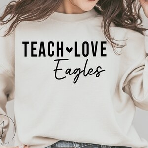Può includere: Una felpa bianca con la scritta "TEACH LOVE Eagles" stampata in nero.
