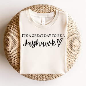 Puede incluir: Una camiseta blanca con el texto "It's a great day to be a Jayhawk" y un símbolo de corazón impreso en ella.