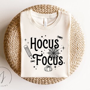 Puede incluir: Una camiseta blanca con el texto "Hocus Focus" en negro. El diseño incluye un lápiz, una telaraña, estrellas y una calabaza.