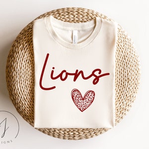 Puede incluir: Una camiseta blanca con la palabra "Lions" en letras rojas y un corazón rojo con un diseño de estampado de leopardo.