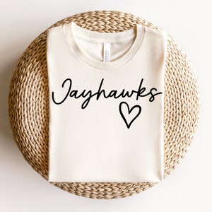 Puede incluir: Una camiseta blanca con el texto "Jayhawks" en letra negra con un pequeño corazón debajo.