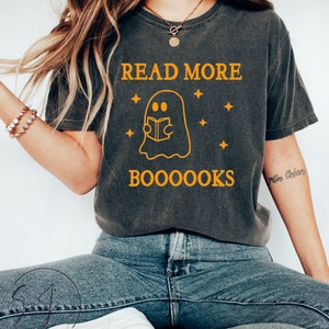 Puede incluir: Una camiseta gris con un gráfico de un fantasma leyendo un libro. El texto "READ MORE BOO0000KS" está impreso en naranja en la camiseta.