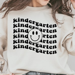 May include: A white sweatshirt with black text that reads "kindergarten" repeated multiple times. A black smiley face is centered in the middle of the text.