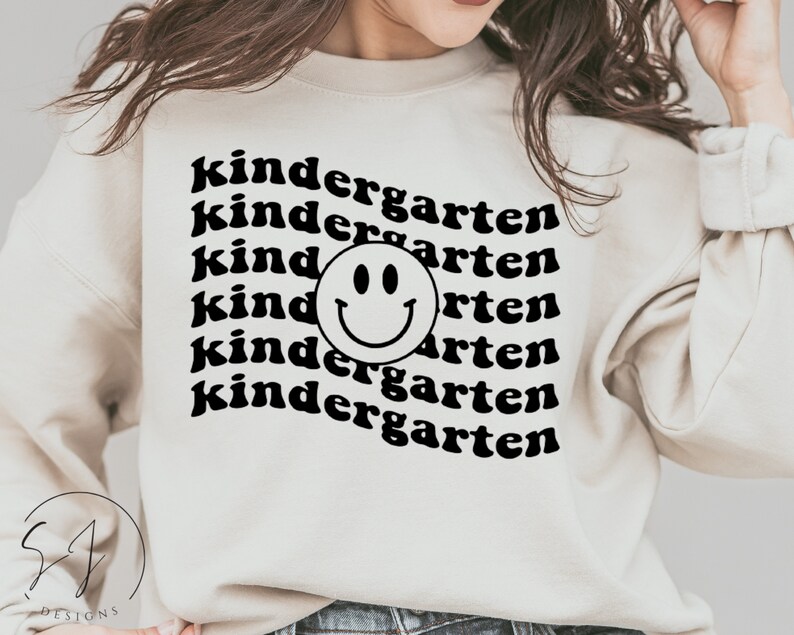 May include: A white sweatshirt with black text that reads "kindergarten" repeated multiple times. A black smiley face is centered in the middle of the text.