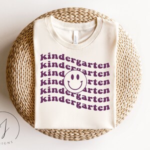 May include: A white t-shirt with the text "kindergarten" repeated multiple times in a purple color. A purple smiley face is centered in the middle of the shirt.