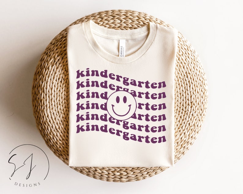 May include: A white t-shirt with the text "kindergarten" repeated multiple times in a purple color. A purple smiley face is centered in the middle of the shirt.