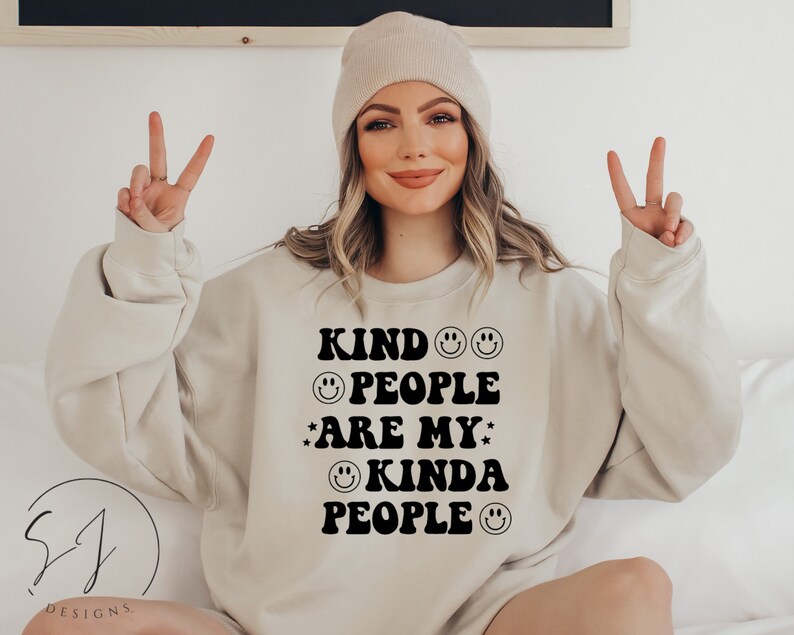 May include: A beige crew neck sweatshirt featuring the phrase "KIND PEOPLE ARE MY KINDA PEOPLE" in black, with smiley faces and stars. The sweatshirt is a neutral colour.