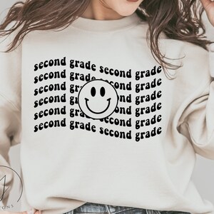 Puede incluir: Una sudadera blanca con el texto "second grade" repetido varias veces en una fuente retro. Una cara sonriente negra está centrada en la sudadera.