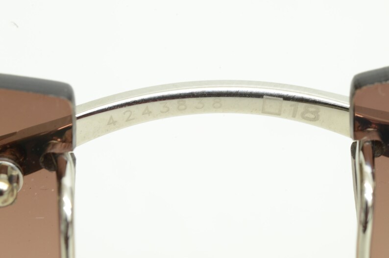 May include: Close-up of the inside of a silver-toned pair of sunglasses with the numbers 4243838 and 18 engraved on the metal arm.