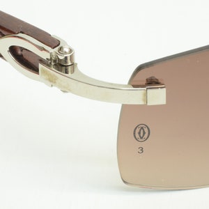 May include: Close-up of the hinge and temple of a pair of brown sunglasses with a silver frame. The sunglasses have a Cartier logo and the number 33 printed on the temple.