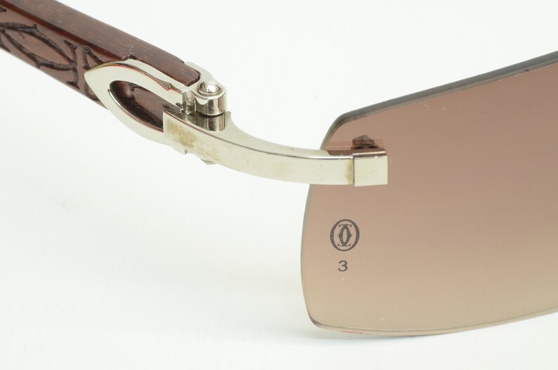 May include: Close-up of the hinge and temple of a pair of brown sunglasses with a silver frame. The sunglasses have a Cartier logo and the number 33 printed on the temple.
