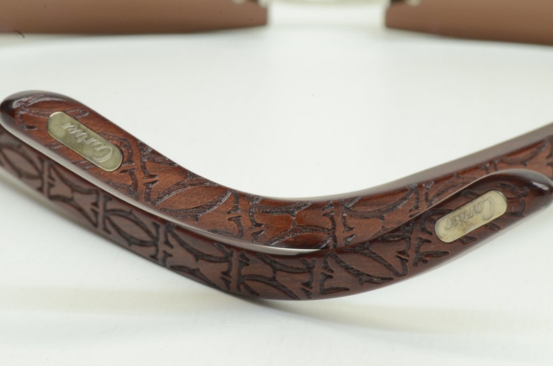 May include: Two brown wooden sunglasses frames with a carved floral design. The frames have a silver metal plate with the word 'Carrera' engraved on them.