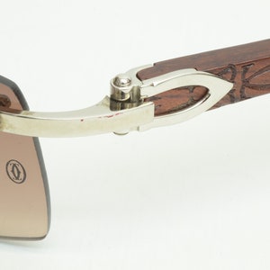 May include: Close-up of the hinge of a pair of brown wood and silver sunglasses. The hinge is silver and has a small, round, silver knob. The sunglasses have a brown wood frame with a carved design. The sunglasses have a brown lens.