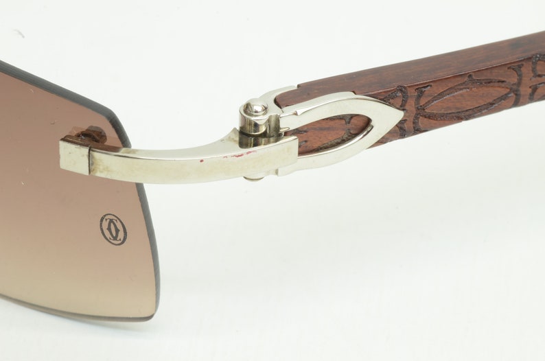 May include: Close-up of the hinge of a pair of brown wood and silver sunglasses. The hinge is silver and has a small, round, silver knob. The sunglasses have a brown wood frame with a carved design. The sunglasses have a brown lens.