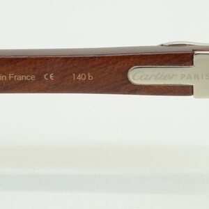 May include: A close-up of the brown wooden arm of a pair of Cartier Paris glasses. The arm has the words 'Made in France', 'CE', '140 b', and 'Cartier Paris' engraved on it.