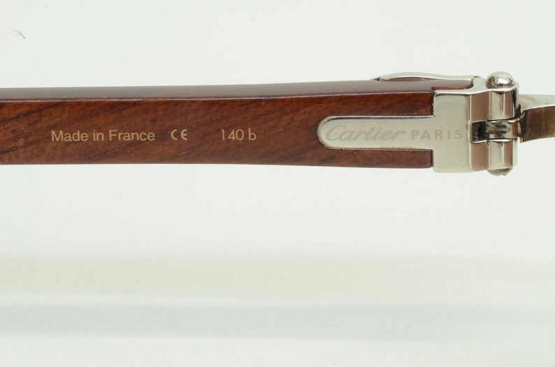 May include: A close-up of the brown wooden arm of a pair of Cartier Paris glasses. The arm has the words 'Made in France', 'CE', '140 b', and 'Cartier Paris' engraved on it.