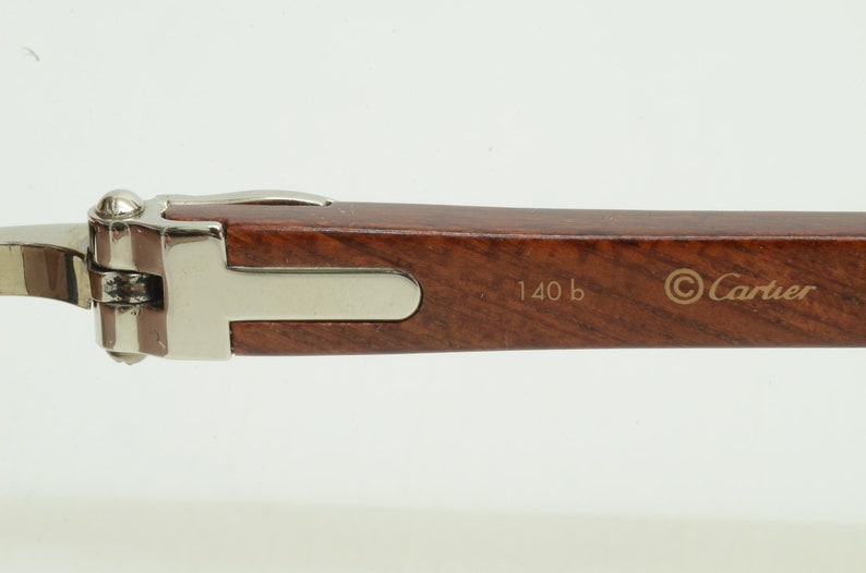 May include: Close-up of the brown wooden arm of a pair of Cartier glasses with silver metal accents. The arm is engraved with the text '140 b' and the Cartier logo.