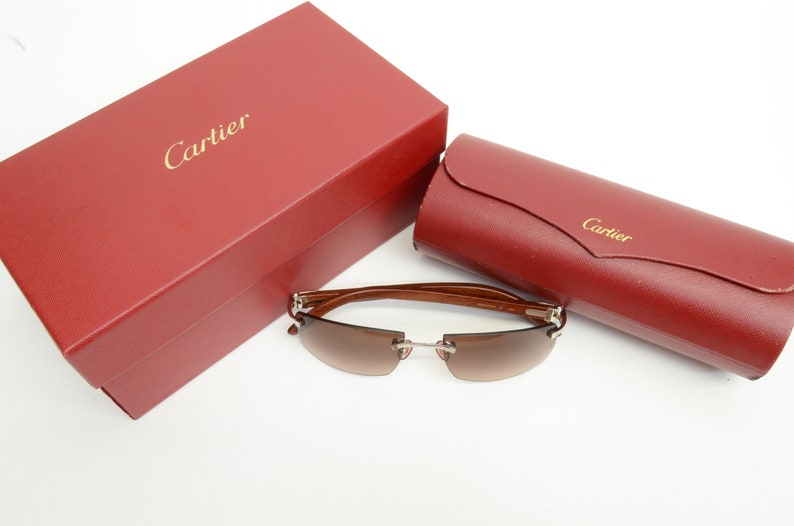 May include: Brown and silver Cartier sunglasses with brown lenses. The sunglasses are resting on a white surface. The sunglasses are next to a red box with the word 'Cartier' on it. The sunglasses are also next to a red case with the word 'Cartier' on it.