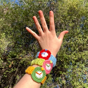 May include: A person's arm wearing four crocheted watch bracelets. The bracelets are in different colours, including red, green, yellow, and pink. Each bracelet has a crocheted clock face.