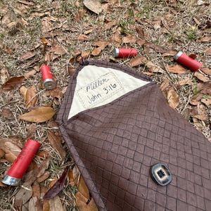 May include: A brown, diamond-patterned case with a button closure, lying on the ground with scattered red shotgun shells. The case has a cream-colored interior with the text "Miller John 3:16".