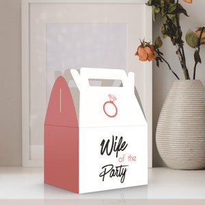 Op de afbeelding: Een witte cadeaudoos met een roze handvat en een roze ringontwerp. Op de voorkant van de doos staat de tekst "Wife of the Party".