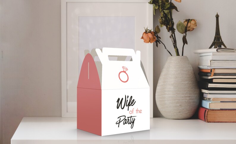 Op de afbeelding: Een witte cadeaudoos met een roze handvat en een roze ringontwerp. Op de voorkant van de doos staat de tekst "Wife of the Party".
