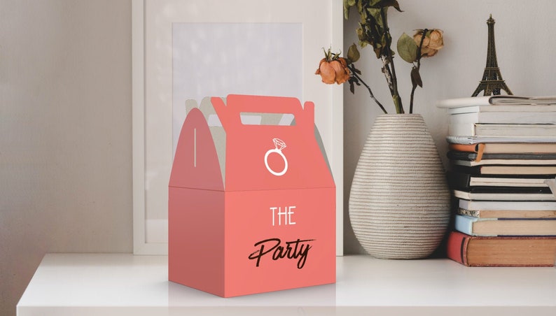 Op de afbeelding: Een roze cadeaudoos met een wit ringontwerp en de tekst "THE Party" op de voorkant. De doos heeft een handvat en is perfect voor kleine cadeautjes of lekkernijen.