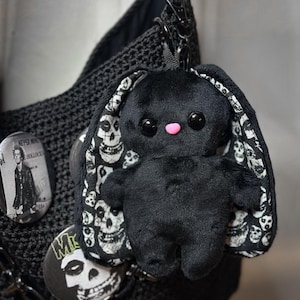 May include: Black plush bunny keychain with skull-patterned ears and a pink nose. The bunny is attached to a black bag with pins featuring band logos. The bag is made of a woven material.