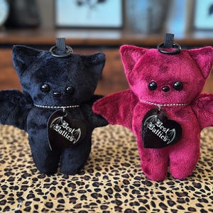 May include: Two plush bat toys, one black and one deep red, each featuring large black eyes, small ears, and outstretched wings. A silver ball chain necklace on each bat holds a black heart-shaped tag with the white text Best Batties.