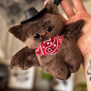 May include: A brown plush bat toy with large black eyes, wearing a small black cowboy hat and a red bandana with a white paisley pattern. The bat has outstretched wings and a black loop for hanging.