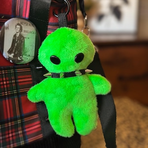 May include: A bright green plush alien toy with black button eyes and a studded collar. The alien is attached to a black strap on a red and black plaid tote bag. A pin with text and a photo is also attached.