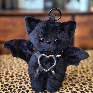 May include: A black plush bat with a heart-shaped harness featuring silver spikes. The bat has large, round eyes, small ears, and attached wings. A black loop is attached to the top of the bat's head.